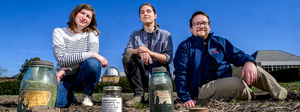 Three researchers sitting in a field with several jars of plant matter